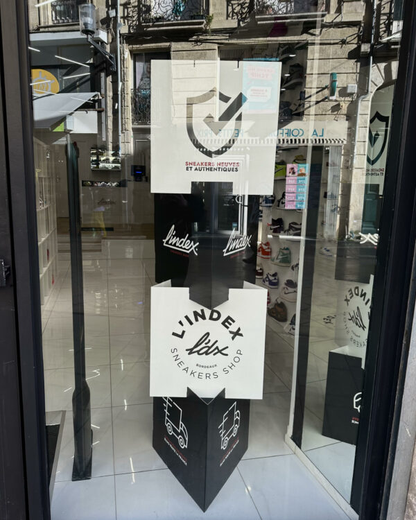 Totem publicitaire noir et blanc de L’Index Shop, visible depuis la vitrine du magasin à Bordeaux.